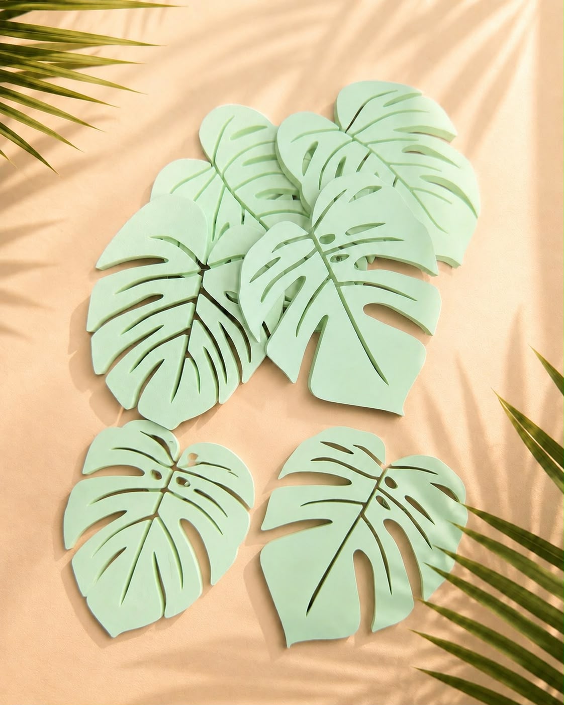 Six sage green Monstera leaf coasters arranged on a soft pink surface