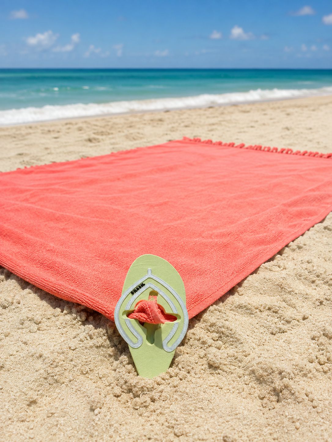 Green flip-flop-shaped beach towel sand anchor securing a coral towel on a sandy beach