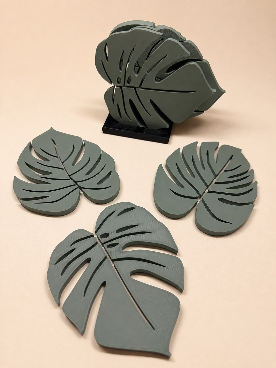 Sage green Monstera leaf coasters with a matching leaf-shaped holder on a soft beige surface